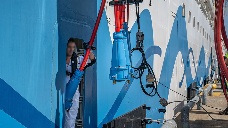 En person i en lucka i ett stort fartyg håller i en stor elkontakt.