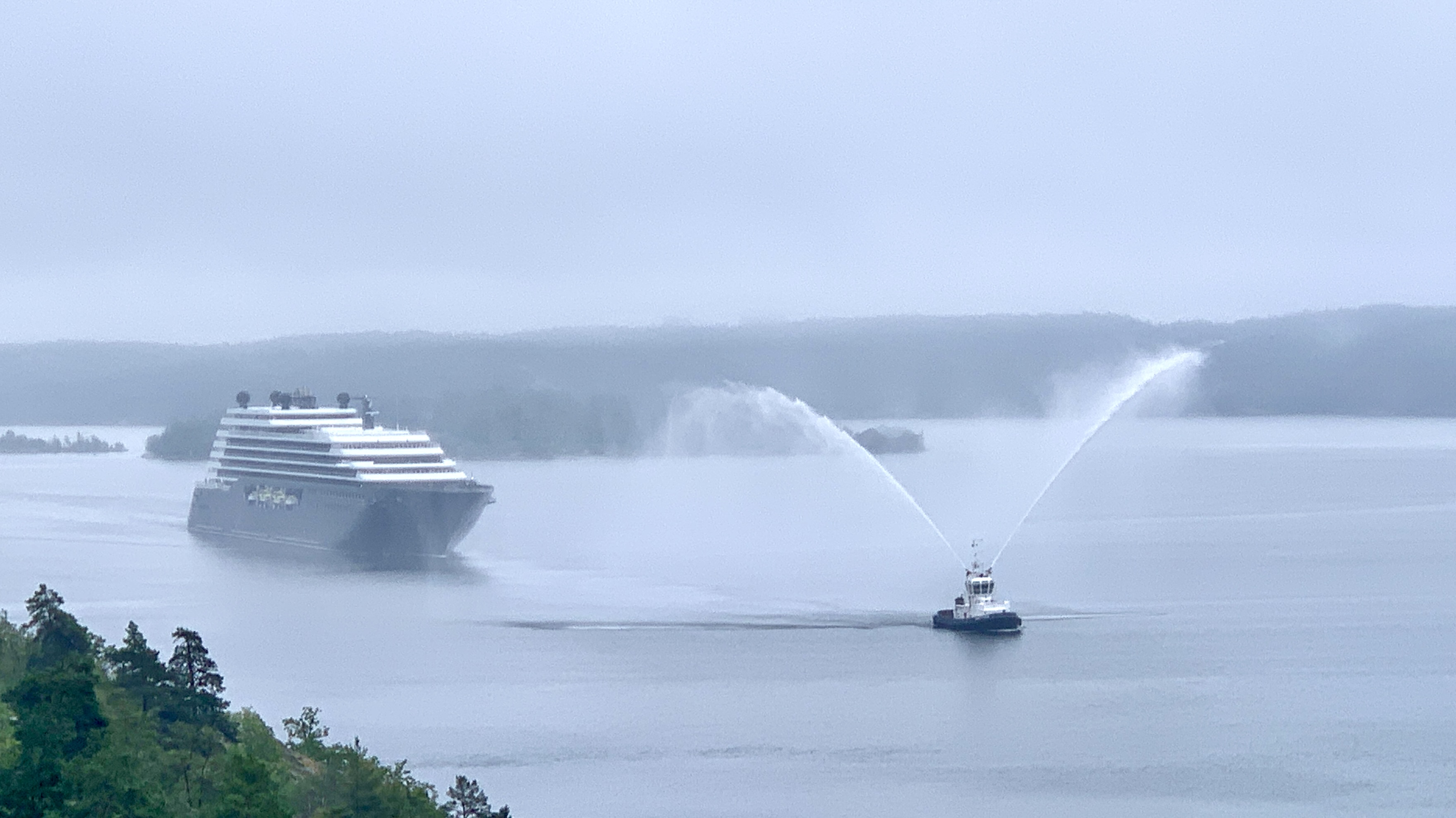 Kryssningsfartyg på väg mot Frihamnen. Framför finns en bogserbåt som sprutar vattensalut.