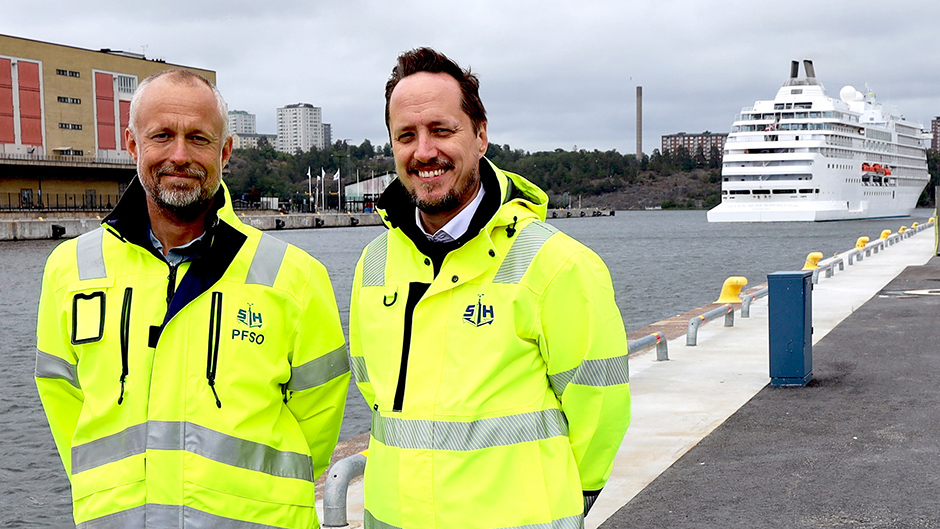 Två män i varseljackor på kajen i Frihamnen. I bakgrunden ett kryssningsfartyg.